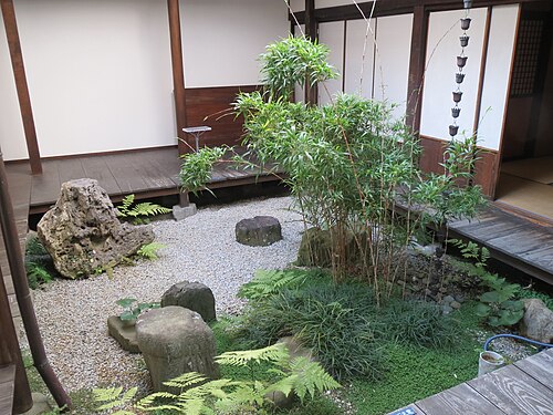 淡い砂利の上に約 30 センチメートル (12 インチ) の縁があり、特徴的な石、雨樋があり、竹、シダ、草、小さな丸い葉を持つつる植物が植えられている中庭。