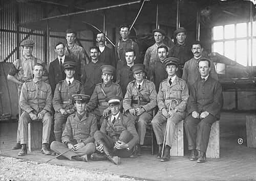 格納庫内の複葉機の前で、軍服に制帽をかぶった6人を含む19人の男性の集合写真。