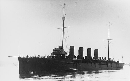 海岸線を背景に、巡洋艦が水上に静かに停泊しています。船の中央には 2 本のマストと 4 本の煙突が目立つように設置されています。