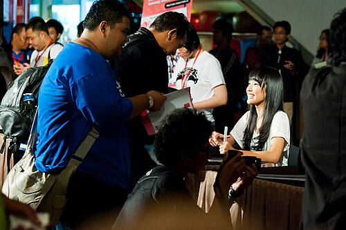 JKT48のメンバーが、サイン会中にファンを見上げて微笑んでいる。