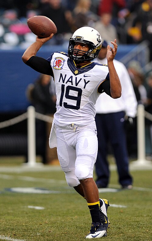 前面に「Navy」と書かれた白いユニフォームを着たアメリカンフットボール選手が、フレーム外のレシーバーにボールを投げている。