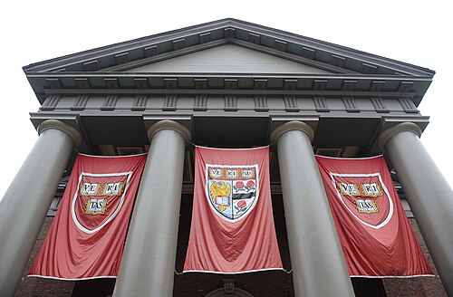 ハーバード大学の壮大な建物。ギリシャ風の 4 本の柱があり、その間に 3 本の赤い旗が垂れ下がっています。t=外側の 2 本の旗には veritas という文字が書かれた紋章が描かれており、中央の旗には veritas という文字に加えて 2 本のバラと本を持った鷲が描かれた紋章が描かれています。