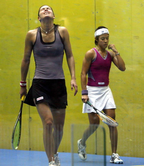 青みがかったトップスを着た女性スカッシュ選手が、紫色のトップスと白いヘッドバンドを着た別のスカッシュ選手が通り過ぎるのをイライラして頭を後ろに倒している。