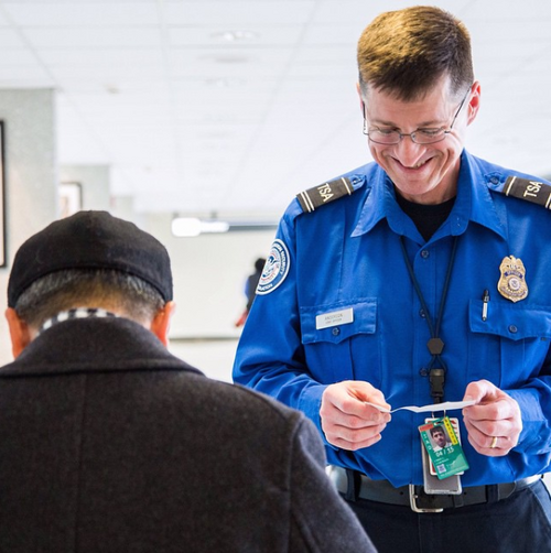 TSA 職員が笑顔で乗客の身分証明書を見下ろしている。乗客は手前にいてカメラから背を向けており、黒い服を着ている。