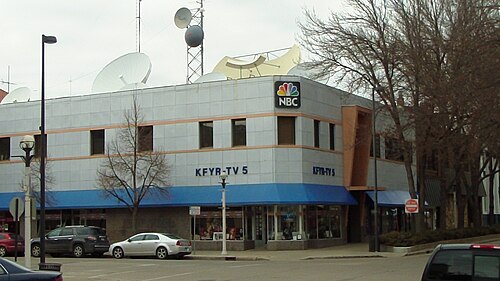 NBC と KFYR-TV の看板がある 2 階建ての建物。屋根には衛星放送受信アンテナとマイクロ波アンテナがいくつか見えます。1 階には他の店舗が入っています。