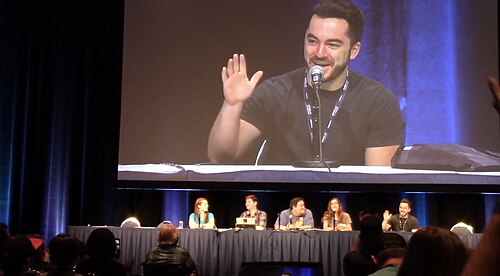 ステージ上には複数のパネリストがおり、そのうちの一人の男性が彼らの頭上の大型スクリーンに映し出されている。