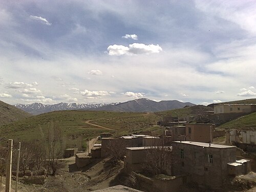 イランの丘陵地にある村を広角で捉えた写真。斜面に建てられた平屋根の石造りと日干しレンガ造りの家々が立ち並んでいる。建物は密集しており、軒下には木製の梁が見える箇所もある。周囲の風景は、澄み切った空の下、乾燥した山々が広がっている。