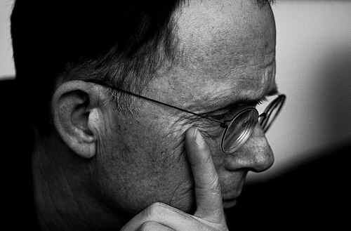眼鏡をかけた中年の白人男性の横顔。彼は右を向いており、左手の人差し指は目の少し下の右頬に置かれています。
