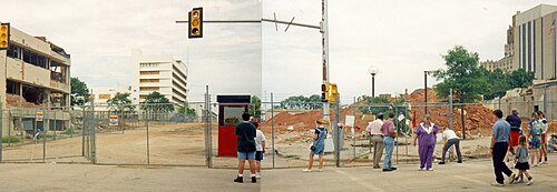 2 枚の画像をつなぎ合わせたもので、建物が取り壊される前の場所を示しています。敷地への入り口を塞ぐ金網フェンスの前には、大勢の人が集まっています。敷地には大きな土の山があり、近くの建物にも被害が見られます。