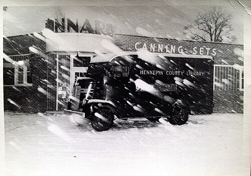 外壁に「CANNING SETS」と書かれたレンガ造りの平屋建ての建物の前に、丸いフロントのトラックが雪の中に停まっている白黒写真。