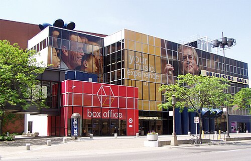 モダンな建物。黄色と青の区画の前に「BOX OFFICE」と書かれた赤い入り口があります。正面には木が植えられています。建物には広告として人の頭の絵が描かれています。