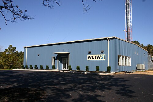A one-story blue metal building with a W L I W logo next to the base of a tall broadcasting tower. In comparison to the prior photo, the building has been reshaped in the front and is larger, with new landscaping.
