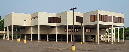 白い3階建ての建物。2階と3階の一部は円筒形の支柱で駐車場の上に吊り下げられている。