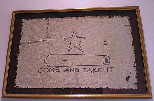 白い旗。小さな大砲の輪郭の上に五芒星が描かれ、その下に「Come and Take It」の文字が書かれている。旗の左隅は引きちぎられ、縫い直されている。
