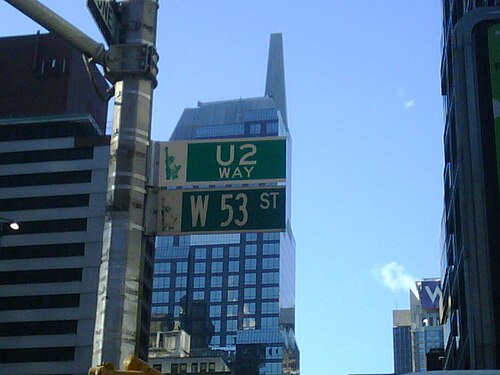 53 番街と書かれた緑色の道路標識。その標識のすぐ上には、同じ色で「U2 Way」と書かれた別の標識があります。反射窓のある高層ビルが標識のすぐ後ろにあります。
