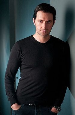 黒いTシャツを着て壁にもたれかかっている、真面目そうな表情の30代後半の黒髪の男性