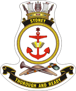 船のバッジ。金色で「SYDNEY」と書かれた黒い巻物の上に海軍の王冠が乗っています。これは、赤い錨が中央にある、ロープ模様の黄色いリングの上にあります。リングの下には、石斧とブーメランの上に座っているヌラヌラがあります。バッジの下部には、「THOROUGH AND READY」と書かれた黒い巻物があります。