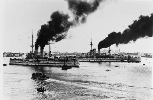 2 隻の戦艦が、周囲に数隻の小型船を従えて港に入港しています。大きな戦艦は背景に、小さな戦艦は前景にあります。