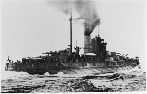 大型戦艦が高速で水上を進んでいます。手前には船尾が見え、船の側面には水が打ち寄せています。煙突からは煙が噴き出しているのが見えます。