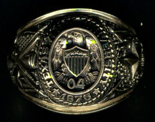 金の指輪のクローズアップ。上部には「Texas A&M University 1876」の文字が、盾の上に鷲が描かれ、その下に数字の0と4が配置されている。左側面には大きな星と樫の木が、右側面には大砲、サーベル、ライフルが描かれ、その下にアメリカ合衆国とテキサス州の国旗が交差して描かれている。