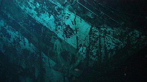 船首の一部のクローズアップ写真。船首は斜めになっています。画像の中央に錨があります。色のついた帯が左上隅から下端の右 3 分の 1 まで伸びています。暗い四角と数字の「7」の残骸が錨と帯の上と左側にあります。船首は錆と海藻で覆われています。画像は深い水中で撮影されたため、青緑色がかっています。