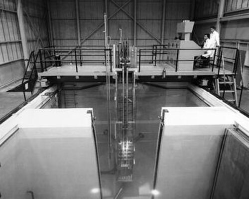 部分的に壁で区切られたプールの白黒画像。プールの後ろには金属製の橋があり、そこから長い金属部品がプールに垂れ下がっています。左側の橋の上で、白衣を着た 2 人の男性がコンソールを見ています。