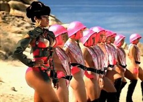 砂漠の岩の前に立つ女性が、ヘルメットをかぶった他の女性たちを率いて砂の上を歩いている。