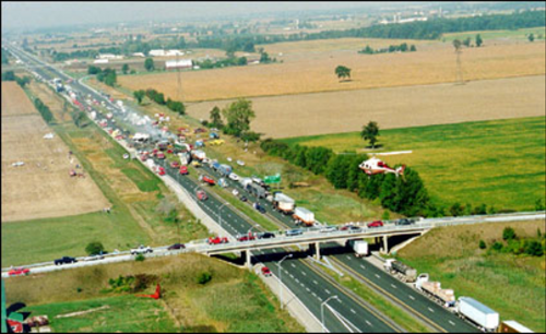 「上空から見た高速道路が、右下から左上にかけて遠くまで伸びています。画像の下部付近では、高架道路によって道路が高速道路を横切っています。周囲は完全に農地です。高速道路では、数十台の車が重なり合っています。大きな玉突き事故の真ん中では煙が上がっています。」