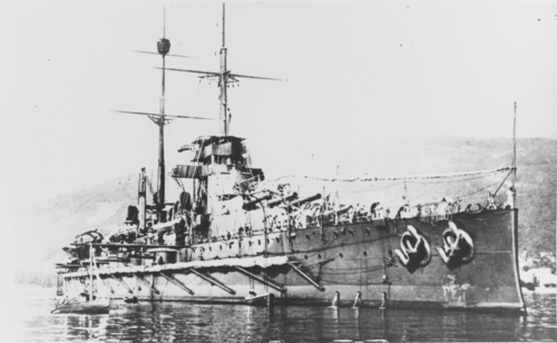 大きな戦艦が陸地近くの水上に静止しています。手前には数隻の小船が見え、背景には戦艦の背後に大きな丘が見えます。