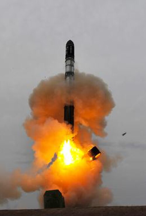オレンジ色の塵と煙の雲の中を地面の穴から登るロケット。ロケットの底から金属のリングが落ちている。