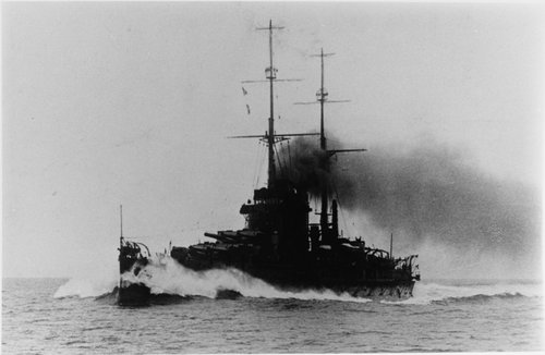 大型戦艦が高速で水上を航行しています。船首に水がぶつかり、煙突から煙が噴き出しています。