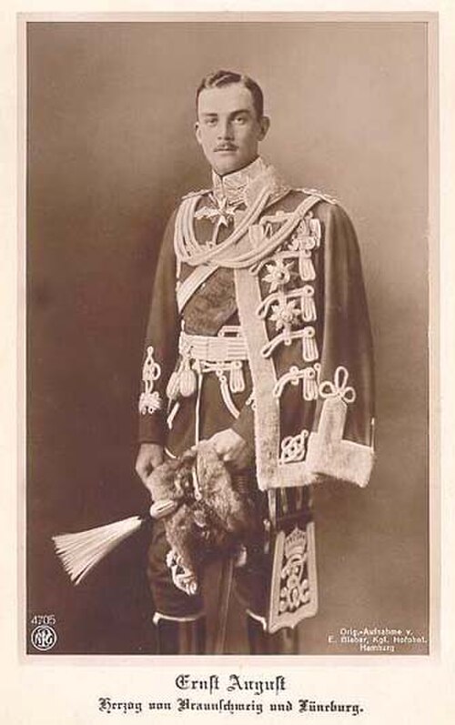 1913年から1918年の在位期間中に撮影された、装飾が施された華やかな軍服を着用し、羽根飾りのついた帽子を手に持った、ブラウンシュヴァイク＝リューネブルク公エルンスト・アウグストの白黒肖像写真。